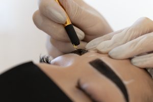 A Microblading artist is using a manual tool on a woman to create fine hair-like strokes for fuller brows.