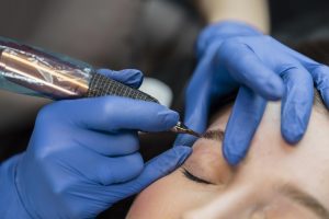 A cosmetic tattoo artist applies pigment to a woman's eyebrow.