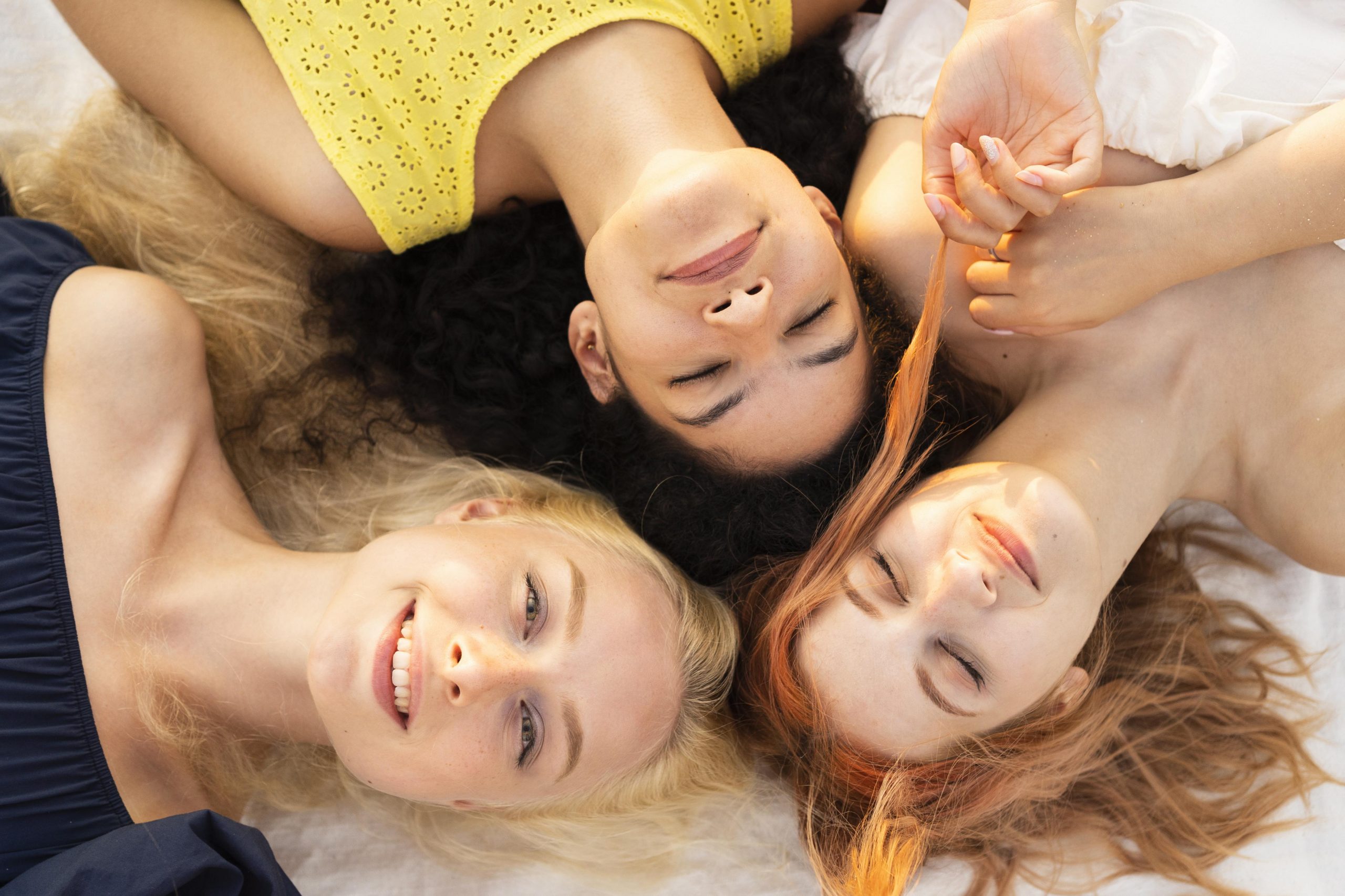 Aerial view of three young women lying down with different coloured eyebrows that match the colour of their hair.