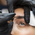 An eyebrow technician using a tool to apply pigment on woman's brows.