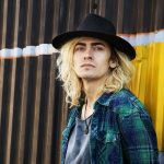 A male blond long-haired rock star stands with his back to a wall. He's endorsing Microblading.